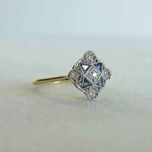 Peut inclure: Une bague en or avec une tête en forme de losange, ornée d'un diamant rond central, de quatre saphirs bleus triangulaires et de quatre petits diamants ronds, le tout serti dans une monture en métal argenté avec des détails milgrain.
