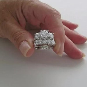 May include: A silver engagement ring set with multiple bands. The rings feature a large, round, clear gemstone as the centerpiece, surrounded by smaller stones. The rings are held in a hand against a white background.