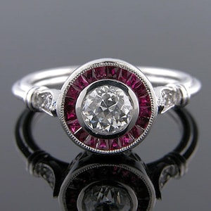 Peut inclure: Une bague en argent avec un diamant rond central entouré d'un halo de rubis rouges. La bande est décorée de petits diamants.
