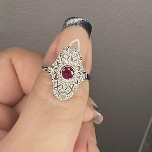 Peut inclure: Bague en argent ornée d'un rubis central et de pierres claires, entourée d'un motif filigrané élaboré. La bague présente un design allongé et détaillé, d'un style vintage ou antique.