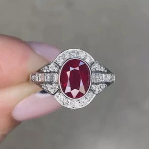 May include: A silver ring with a central oval, deep red gemstone, encircled by smaller, clear stones. The ring has an intricate design with rectangular stones on the sides. The ring's design suggests a vintage style. The ring is held up by a hand.