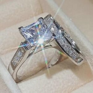 May include: A silver engagement ring set with a large, square-cut clear gemstone. The band is embellished with smaller, sparkling stones. The set includes a matching band, creating a coordinated and elegant appearance.