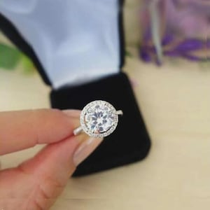 May include: A silver ring with a large, round, clear gemstone at the center, surrounded by a halo of smaller stones. The ring is held in front of a black ring box.