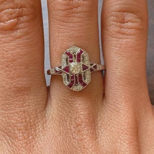 Peut inclure: Bague en argent ornée d'une pierre centrale claire et ronde, entourée de petites pierres claires et de pierres rouges. La bague présente un design Art Déco avec des formes géométriques, portée au doigt.