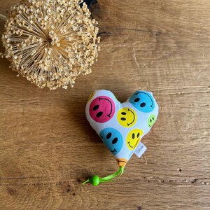 Könnte beinhalten: Ein weißes herzförmiges Kissen mit einem grünen Band und einer grünen Perle. Das Kissen ist mit einem Muster aus bunten Smiley-Gesichtern bedeckt.