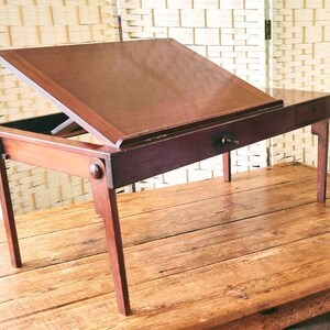 May include: A dark wood writing desk with a hinged top and a drawer. The desk has turned legs and is sitting on a wooden surface.
