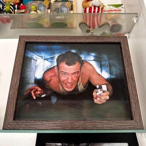 May include: A framed photograph of a man with short brown hair and a goatee, wearing a white tank top, looking at the camera with a serious expression. He is holding a lighter in his right hand. The background is a dark blue and gray.