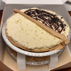 May include: Two pies in a cardboard box, one key lime and one chocolate. The key lime pie has a light green topping, while the chocolate pie has a dark chocolate drizzle and cookie crumble. Both are in foil pie tins.