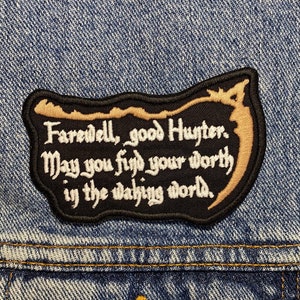 Op de afbeelding: Een geborduurde patch in zwart en goud met de tekst "Farewell, good Hunter. May you find your worth in the waking world."