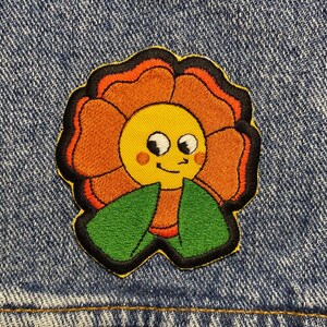 Pode incluir: Uma flor laranja sorridente com folhas verdes, bordada em uma jaqueta jeans azul.