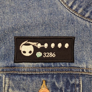 Peut inclure: Patch brodé noir et blanc avec un fantôme de dessin animé tenant un joyau bleu et le numéro 3286. Le patch est cousu sur une veste en jean bleue.