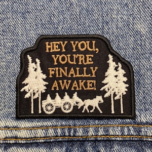 May include: Black and white embroidered patch with the text "Hey you, you're finally awake!" on a black background. The patch features a wagon pulled by two horses and two pine trees on either side.