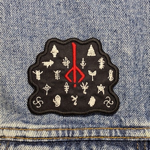 May include: A black and red embroidered patch featuring a symbol surrounded by other symbols. The patch is sewn onto a blue denim jacket.