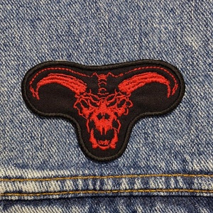 Può includere: Toppa ricamata rossa di un demone cornuto su sfondo nero, cucita su una giacca di jeans blu.