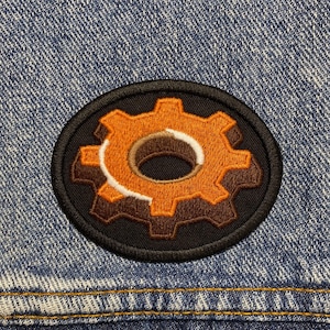 May include: A black and orange embroidered patch of a gear with a brown center.