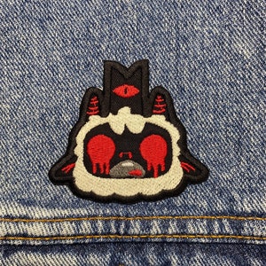 May include: A black and white embroidered patch of a cartoon creature with red eyes and a single red eye on its head. The creature has a white fluffy body and black horns. The patch is sewn onto a blue denim jacket.