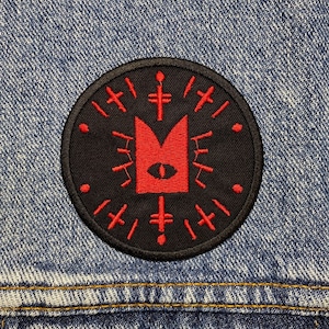 May include: A black and red embroidered patch with a stylized eye and a geometric design. The patch is circular and has a black border.