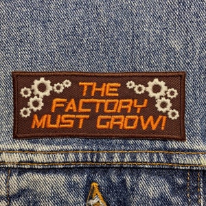 May include: A brown denim jacket with a brown patch that says "THE FACTORY MUST GROW!" with white gears around the text.