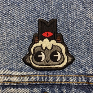 May include: A black and white embroidered patch of a cartoon character with a single red eye, a black hat, and a white fluffy head. The patch is sewn onto a blue denim jacket.