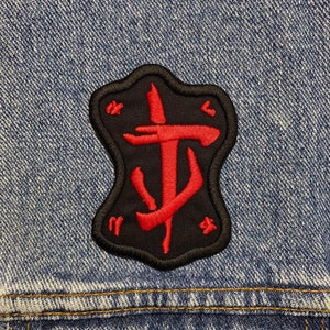 Peut inclure: Patch brodé rouge et noir avec un symbole stylisé ressemblant à un trident ou une lance à trois pointes, sur un fond en jean.