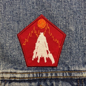 Op de afbeelding: Een rode, vijfhoekige patch met een witte silhouet van een figuur met wapens, tegen een bergketen en een zon. Genaaid op een blauw spijkerjasje.