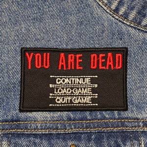 Puede incluir: Un parche bordado negro y rojo con el texto "YOU ARE DEAD" y las opciones "CONTINUE", "LOAD GAME" y "QUIT GAME" en una chaqueta vaquera azul.