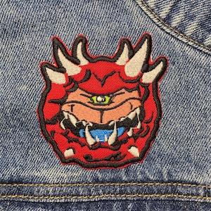 May include: A red and white embroidered patch of a cartoon monster with a single eye and sharp teeth, sewn onto a blue denim jacket.