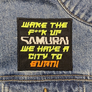 Könnte beinhalten: Ein schwarzer Patch mit weißem und gelbem Text, der "Wake the F**k Up Samurai We Have A City To Burn" lautet. Der Patch ist auf eine blaue Jeansjacke genäht.