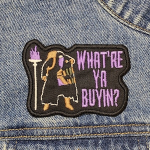 May include: A black and purple embroidered patch with a cartoon figure holding a staff and a purple banner that reads "What're Ya Buyin?"