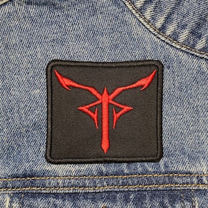 May include: A red and black embroidered patch with a stylised design. The patch is sewn onto a blue denim jacket.