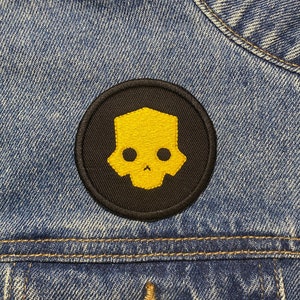 Pode incluir: Um remendo de caveira preto e amarelo com contorno amarelo. O remendo é costurado em uma jaqueta jeans azul.