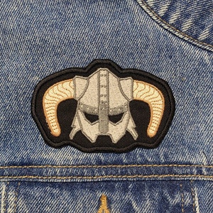 Może przedstawiać: Naszywkę haftowaną w kolorze czarno-białym przedstawiającą hełm z rogami na niebieskiej dżinsowej kurtce. Hełm jest w stylu nordyckim z maską na twarz.