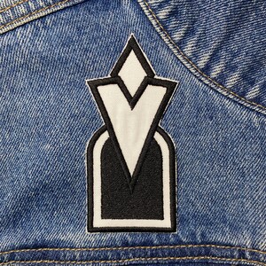 May include: White and black embroidered patch with a geometric design. The patch is sewn onto a blue denim jacket.