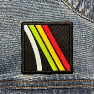 Könnte beinhalten: Ein quadratischer, gestickter Aufnäher mit schwarzem Hintergrund und einem Design aus vier diagonalen Streifen in Weiß, Grün, Gelb und Rot. Der Aufnäher ist auf eine blaue Jeansjacke genäht.