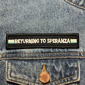 Könnte beinhalten: Nahaufnahme einer Jeansjacke mit einem schwarzen Aufnäher mit dem weißen Schriftzug "RETURNING TO SPERANZA". Der Aufnäher hat einen hellblauen und grünen Streifen. Ein kupferfarbener Knopf ist auf der Tasche zu sehen.