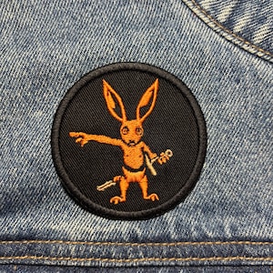 Könnte beinhalten: Ein runder, schwarzer, gestickter Aufnäher mit einer orangefarbenen, kaninchenartigen Figur, die ein Schwert hält und zeigt. Der Aufnäher ist auf eine Jeansjacke genäht. Das Kaninchen hat lange Ohren und ist in einer dynamischen Pose.