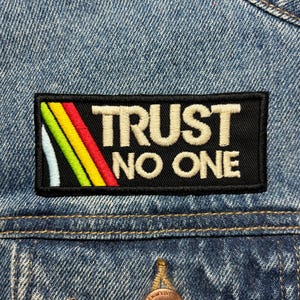 以下が含まれることがあります： ベージュの糸で「TRUST NO ONE」と刺繍された黒い長方形のパッチ。パッチには、赤、黄、緑、水色のストライプのカラフルなストライプデザインがあります。パッチはデニムジャケットに縫い付けられています。