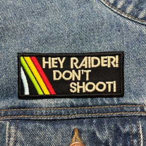 May include: A black rectangular patch with the text "HEY RAIDER! DON'T SHOOT!" in white. The patch has a colourful design on the left side with white, green, yellow, and red stripes. The patch is sewn onto a blue denim jacket.