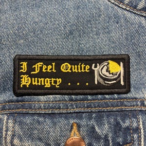 May include: A black rectangular patch with yellow gothic lettering reading "I Feel Quite Hungry". A graphic of a plate with food is on the right. The patch is attached to a blue denim jacket.