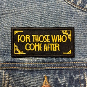 Peut inclure: Un écusson rectangulaire noir avec le texte brodé en or "FOR THOSE WHO COME AFTER". L'écusson présente des motifs d'angle dorés et est cousu sur une veste en jean bleue.
