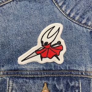 Könnte beinhalten: Ein gestickter roter und weißer Aufnäher des Rittercharakters aus dem Videospiel Hollow Knight, auf eine blaue Jeansjacke genäht.
