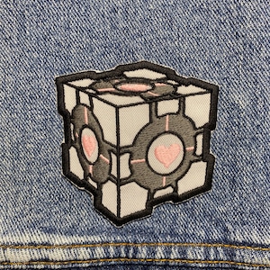 May include: A white and grey embroidered patch of a cube with a heart on each side. The cube is outlined in black and has a grey and white pattern.