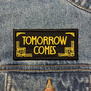 May include: A black rectangular patch with the words "TOMORROW COMES" embroidered in yellow. The patch has yellow decorative corner designs and is sewn onto a denim jacket.