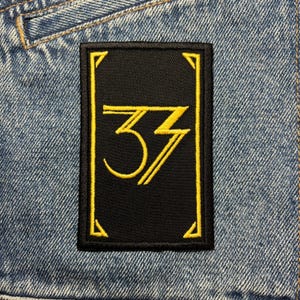 May include: Embroidered black rectangular patch with a gold border and the number "33" in gold. The patch is sewn onto a denim jacket. The number "33" is stylized with a unique font.