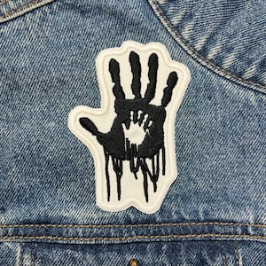 May include: A black handprint patch with a white border, featuring a smaller handprint inside, and dripping black ink design. The patch is sewn onto a blue denim jacket.