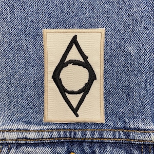 May include: A close-up of a blue denim jacket with a stitched patch. The patch is a cream-coloured rectangle with a black geometric design. The jacket has a button closure and yellow stitching.