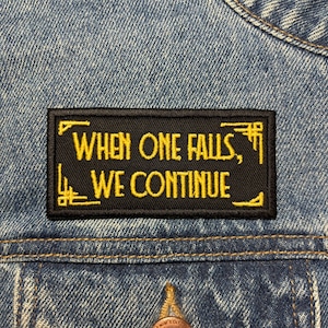May include: A black rectangular patch with yellow embroidered text that reads "WHEN ONE FALLS, WE CONTINUE." The patch is sewn onto a denim jacket. The patch has decorative yellow corner designs.
