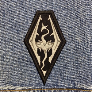 May include: A black and white embroidered patch featuring a dragon with wings outstretched in a diamond shape.