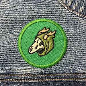 Könnte beinhalten: Gestickter Aufnäher mit einem stilisierten Schädel mit Geweih, der eine grüne Kapuze trägt, auf einem grünen kreisförmigen Hintergrund mit grünem Rand. Der Aufnäher ist auf einer Jeansjacke befestigt und zeigt das Design und die Platzierung.