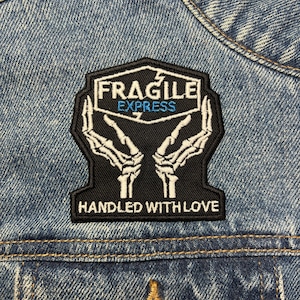 Może przedstawiać: Czarno-biała naszywka z napisem "FRAGILE EXPRESS" i "HANDLED WITH LOVE". Naszywka przedstawia wzór szkieletowych dłoni. Naszywka jest przyszyta do dżinsowej kurtki.